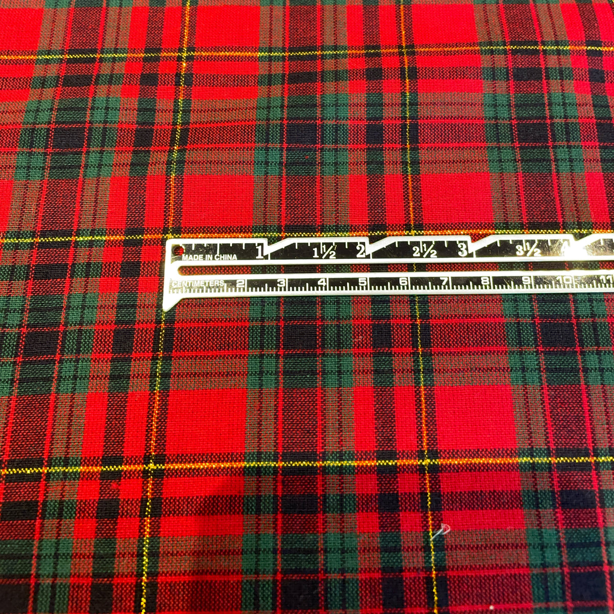 A close-up of red tartan plaid fabric with green, black, and thin yellow stripes, with a metal ruler laid horizontally across the center for scale.