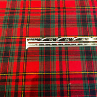 A close-up of red tartan plaid fabric with green, black, and thin yellow stripes, with a metal ruler laid horizontally across the center for scale.