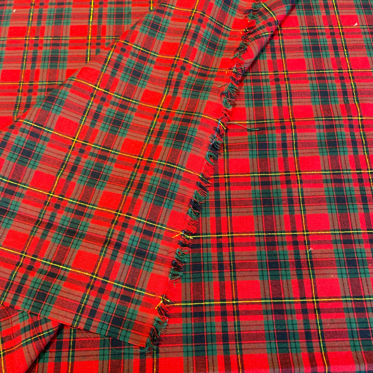 A close-up of red tartan plaid fabric with fringe edges, featuring intersecting black, green, and yellow stripes.