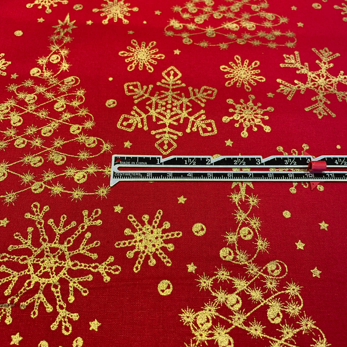 Red fabric with a gold glittery snowflake and Christmas tree pattern, shown with a metal ruler for scale.