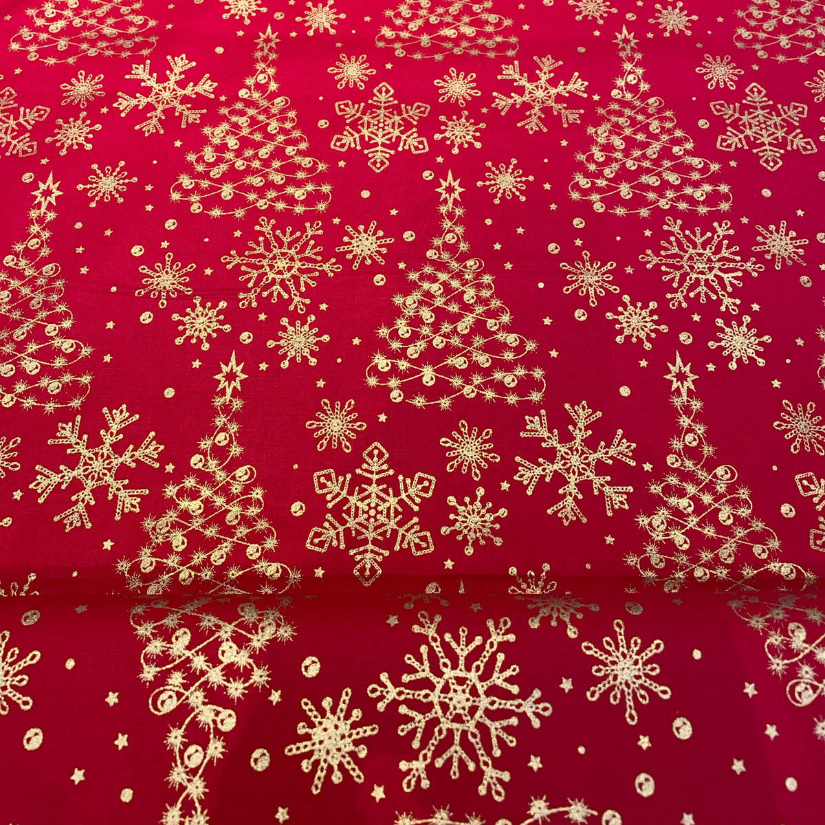 Red fabric with a repeating gold pattern of Christmas trees, snowflakes, and stars, shown folded to reveal the texture and underside.