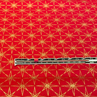 Red fabric with a repeating gold starburst pattern, shown flat with a metal ruler placed horizontally for scale.
