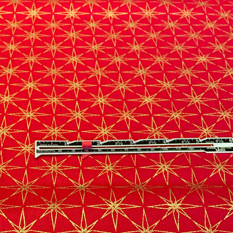 Red fabric with a repeating gold starburst pattern, shown flat with a metal ruler placed horizontally for scale.