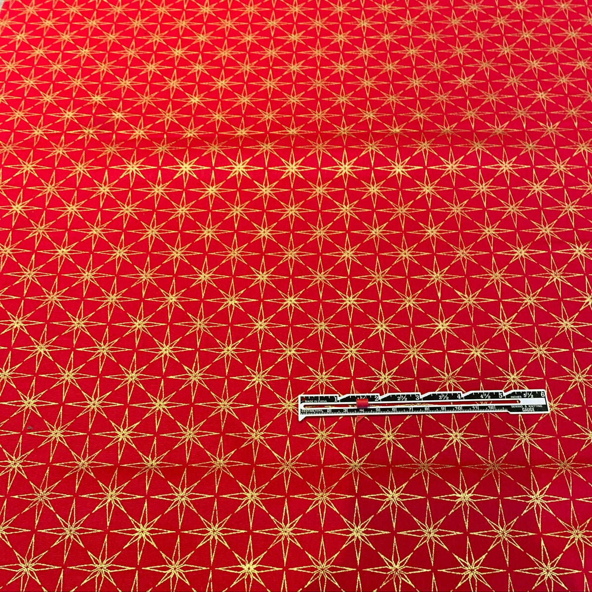 Red fabric with a repeating gold geometric star pattern, shown flat with a small white measurement tape measure placed near the bottom center.