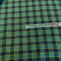 A close-up of a green and black plaid fabric with yellow dashed stitched lines, with a silver metal ruler placed diagonally across the center for scale.