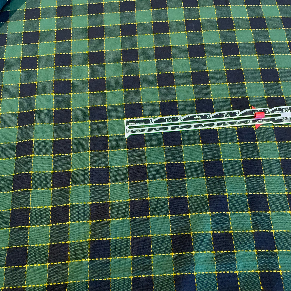 A close-up of a green and black plaid fabric with yellow dashed stitched lines, with a silver metal ruler placed diagonally across the center for scale.