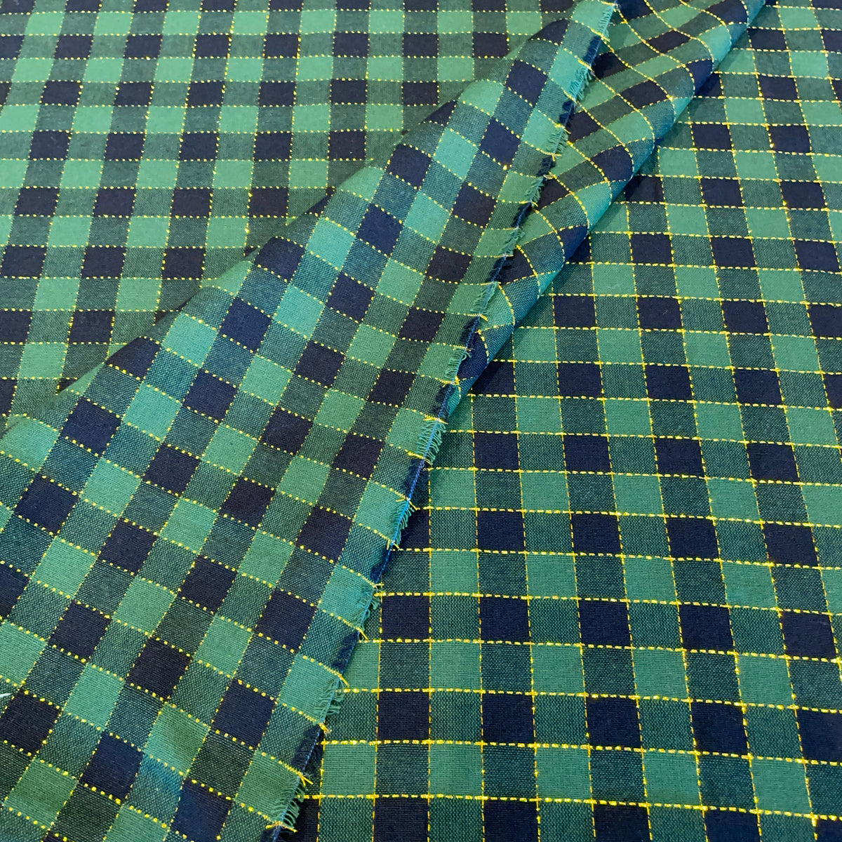 A close-up of plaid fabric with a green and navy blue checkered pattern outlined by thin yellow stitching, folded diagonally across the frame.